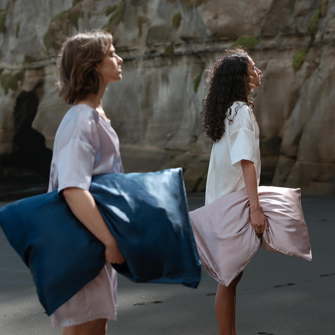 Two women standing on a dark sandy beach near rocky cliffs, each holding a large silk pillow — one navy, one blush. - #color_navy-blue