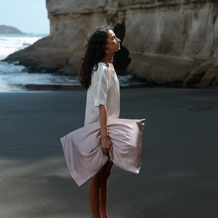 Woman in white dress holding a rose-mist colored silk pillow on a black sand beach - #color_rose-mist
