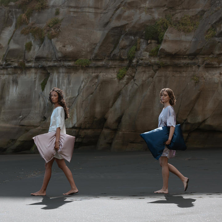 Two barefoot women walking on a dark sandy beach, each holding a large pillow, one pink and one blue, with rocky cliffs behind. - #color_rose-mist