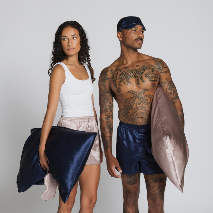 Woman and tattooed man standing side-by-side holding navy and blush silk pillowcases; woman holds navy sleep mask.#color_navy-blue