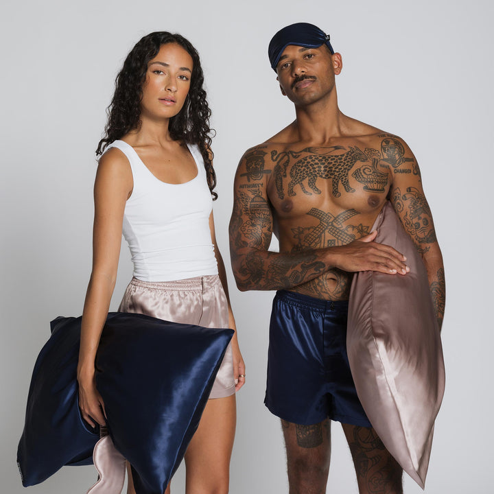 Tattoed man in sleep cap and navy shorts and woman in white tank and silk shorts holding navy and blush silk pillows against gray background. -#color_rose-mist