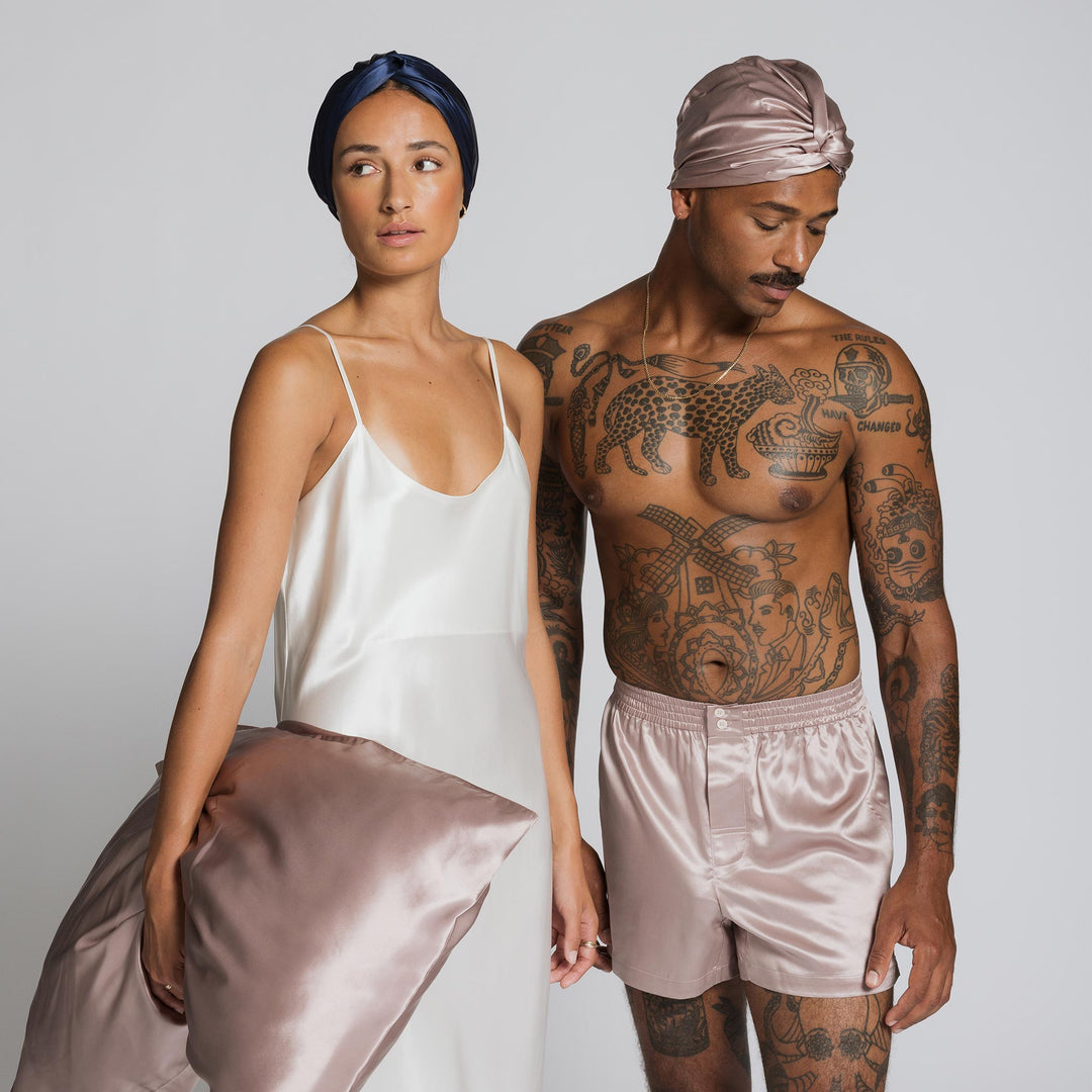 Woman in white silk slip holding a rose-mist silk pillow; man wearing rose-mist silk boxer shorts and turban. - #color_rose-mist