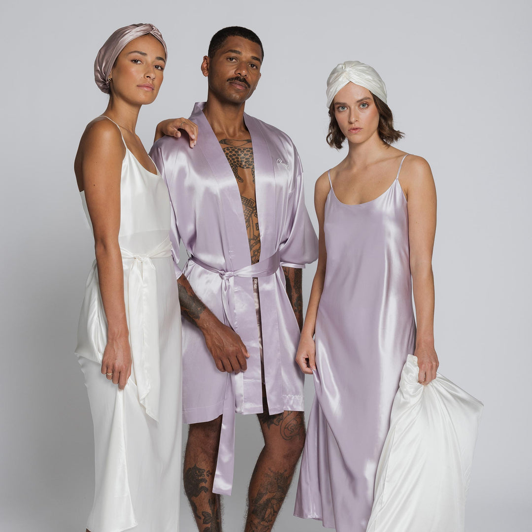 Three models in silk loungewear: woman in white slip with turban; man in lilac robe; woman in lavender slip holding pillow. - #color_pearl-white