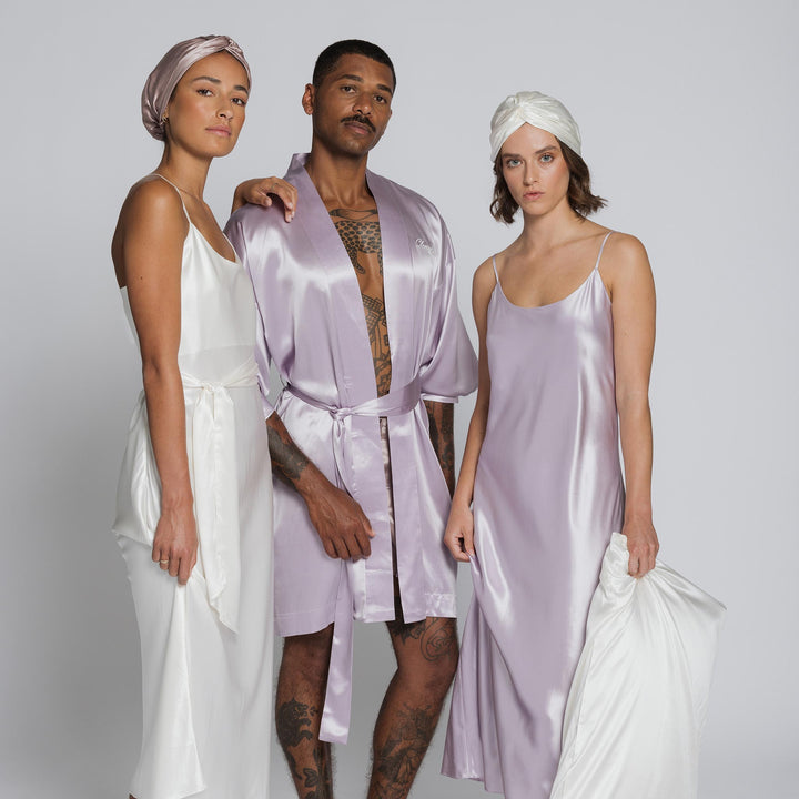 Three models in silk loungewear: woman in white slip with turban; man in lilac robe; woman in lavender slip holding pillow. - #color_pearl-white