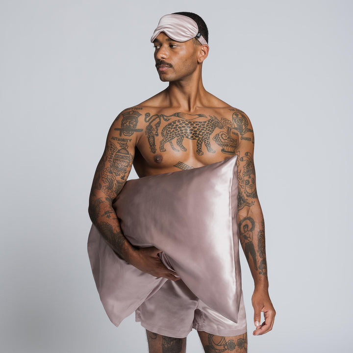 Tattooed man wearing rose-mist silk sleep mask and matching shorts, holding rose-mist silk pillow; tattoo reads AUTHORITY. - #color_rose-mist