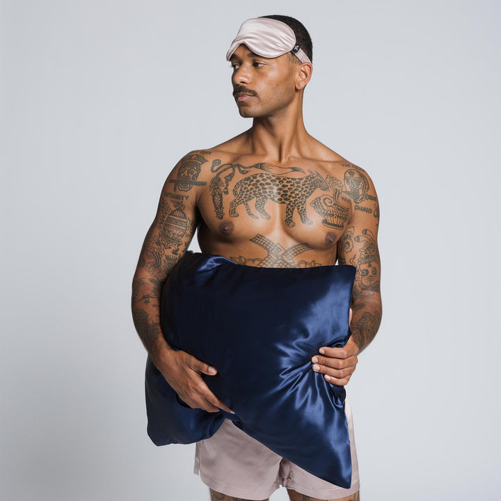 Tattooed man wearing a pink silk sleep mask and holding a navy-blue silk pillow against his chest. - #color_navy-blue