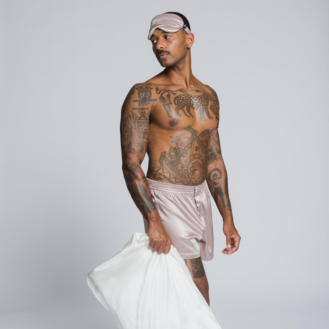 Male model wearing pearl-white silk sleep shorts and mask, holding a white pillow; torso tattoos include the word AUTHORITY. - #color_pearl-white