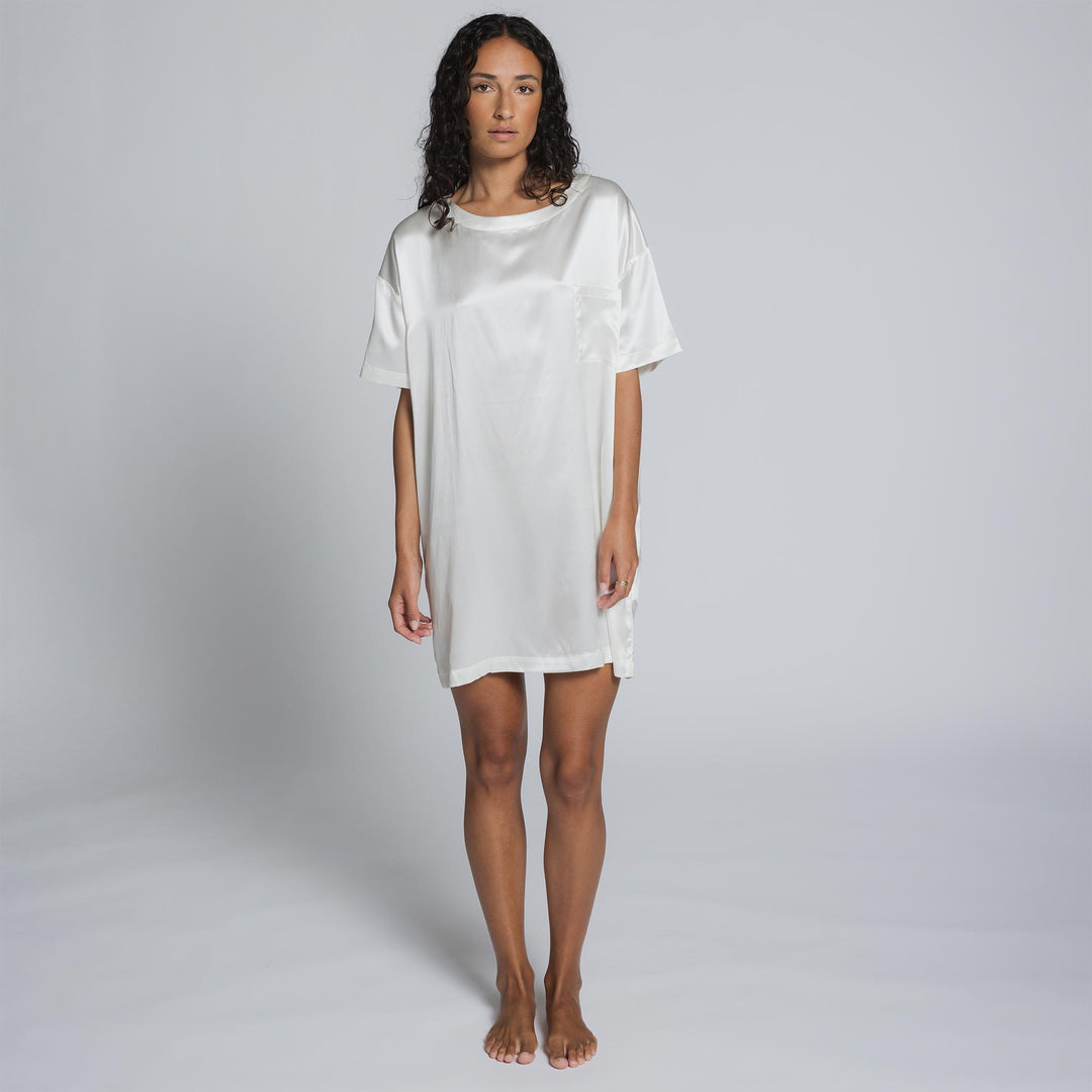 Woman with curly hair wearing a white satin short-sleeve dress, standing barefoot against a plain gray background.