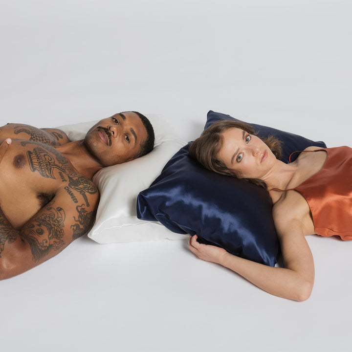 Man with chest tattoos resting on a pearl-white pillow beside a woman lying on a navy silk pillow - #color_pearl-white