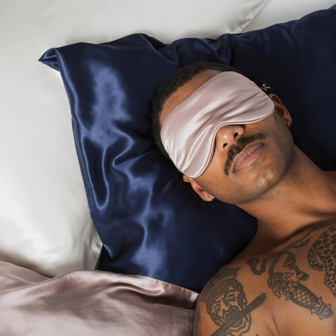 Man with mustache and chest tattoos resting on navy-blue silk pillow, wearing pale-pink silk sleep mask. - #color_navy-blue