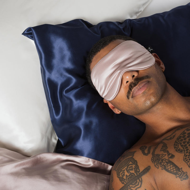 Man with mustache and chest tattoos resting on navy-blue silk pillow, wearing pale-pink silk sleep mask. - #color_navy-blue