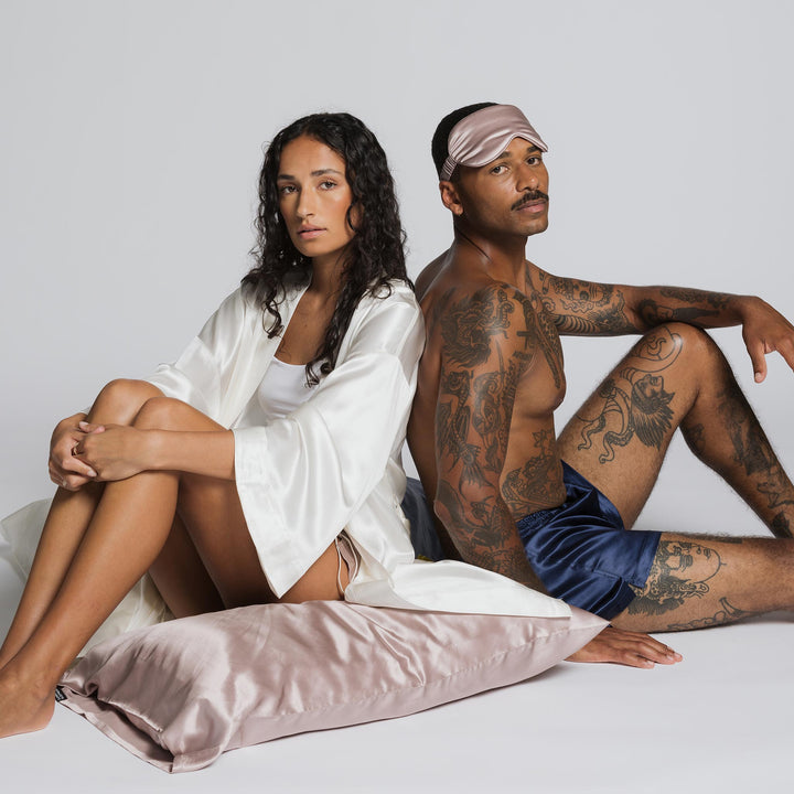 Woman in white silk robe and tattooed man wearing rose-mist sleep mask, seated back-to-back on a rose-mist silk pillow. - #color_rose-mist