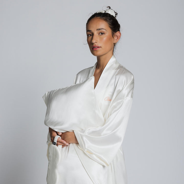 Woman in pearl-white silk robe embroidered 'Clare', holding matching silk pillow against gray background. - #color_pearl-white