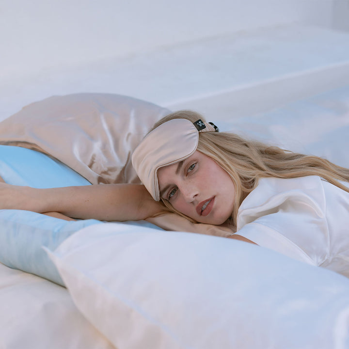 Woman reclining on bed with champagne silk pillowcase and matching silk sleep mask