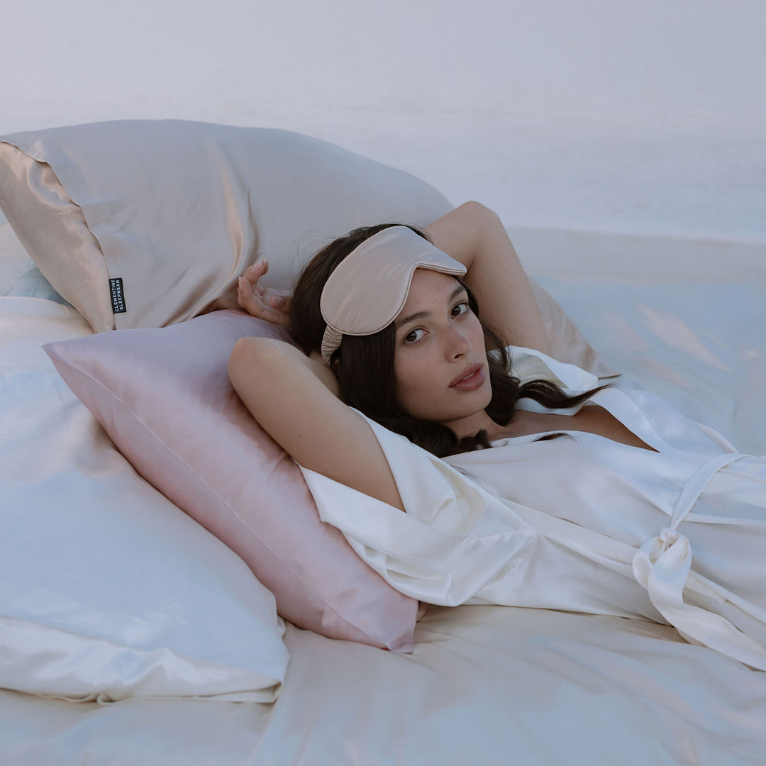 Woman reclining on silk sheets and silk pillows wearing a silk robe and sleep mask pushed above her eyes - laying on bed white pillows#color_pearl-white