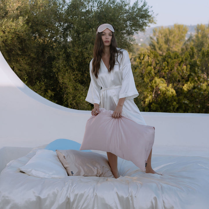 Woman in white robe holding pink pillow standing on bed outdoors with greenery background - #color_pink-sherbert