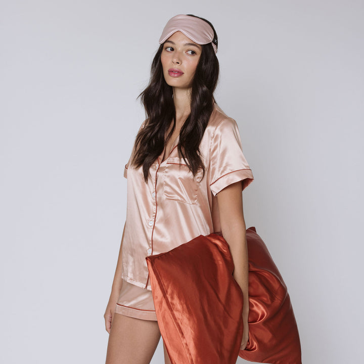 Woman wearing blush satin pajamas and eye mask holding a rust-colored silk pillowcase.#color_dusk