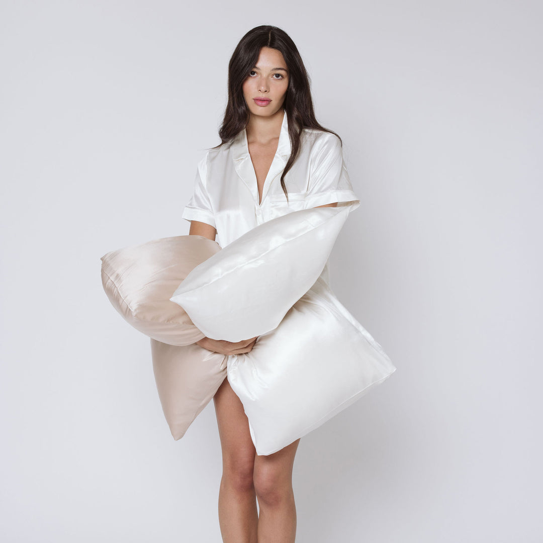Woman in silk pajama shirt holding two silk pillowcases, pearl-white and blush - white pillowcase in hands#color_pearl-white