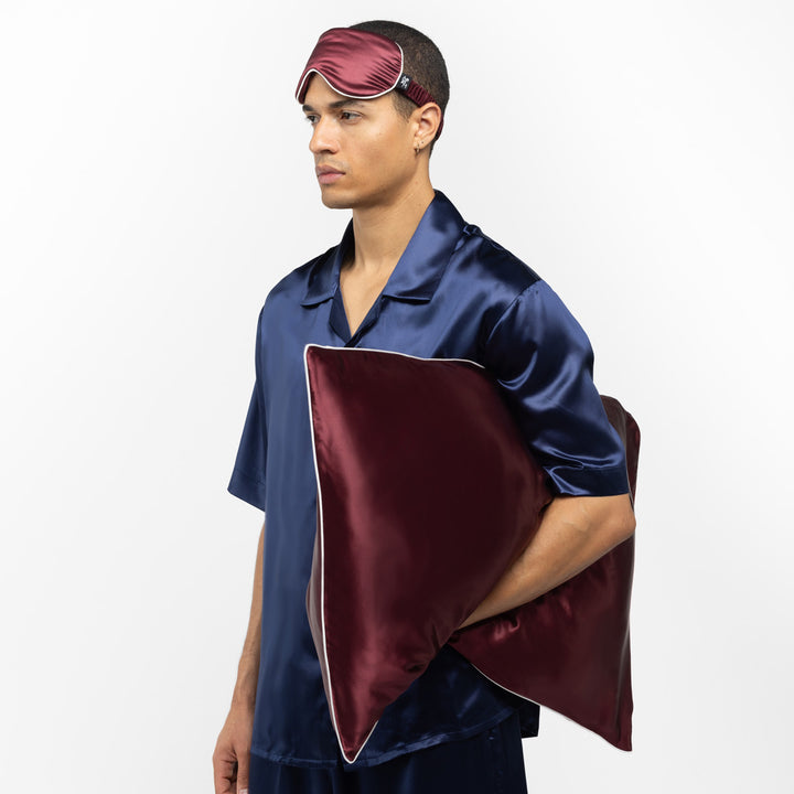 Man in navy silk pajamas wearing a plum silk sleep mask and holding a large plum silk pillow with white piping. - #color_plum