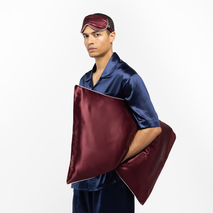 Man in navy silk pajamas holding a plum silk pillow and wearing a matching plum sleep mask - #color_plum