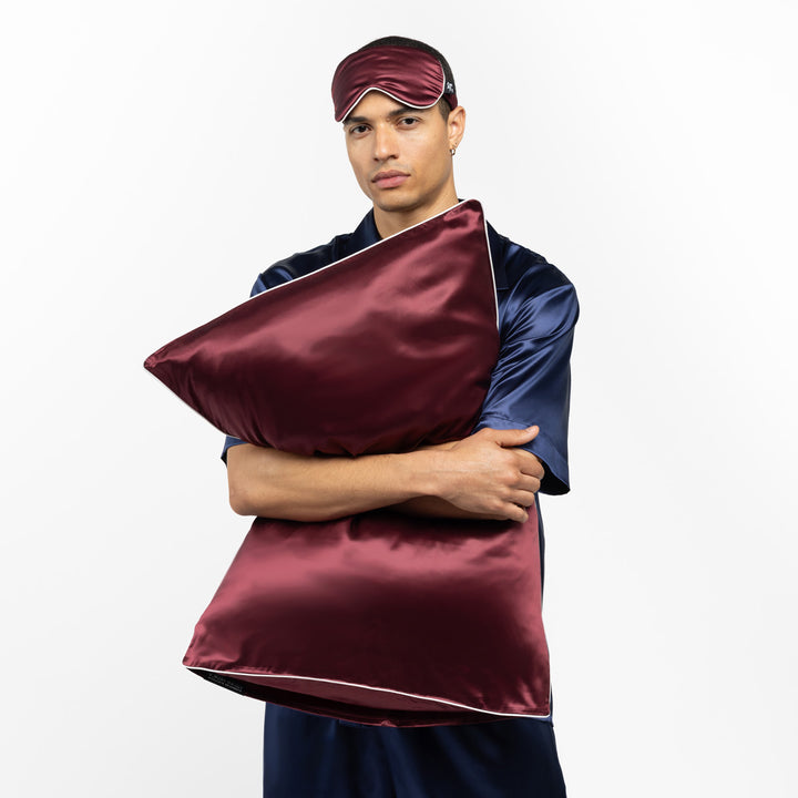Man in navy pajamas holding a plum-colored silk pillow and wearing a matching sleep mask - #color_plum