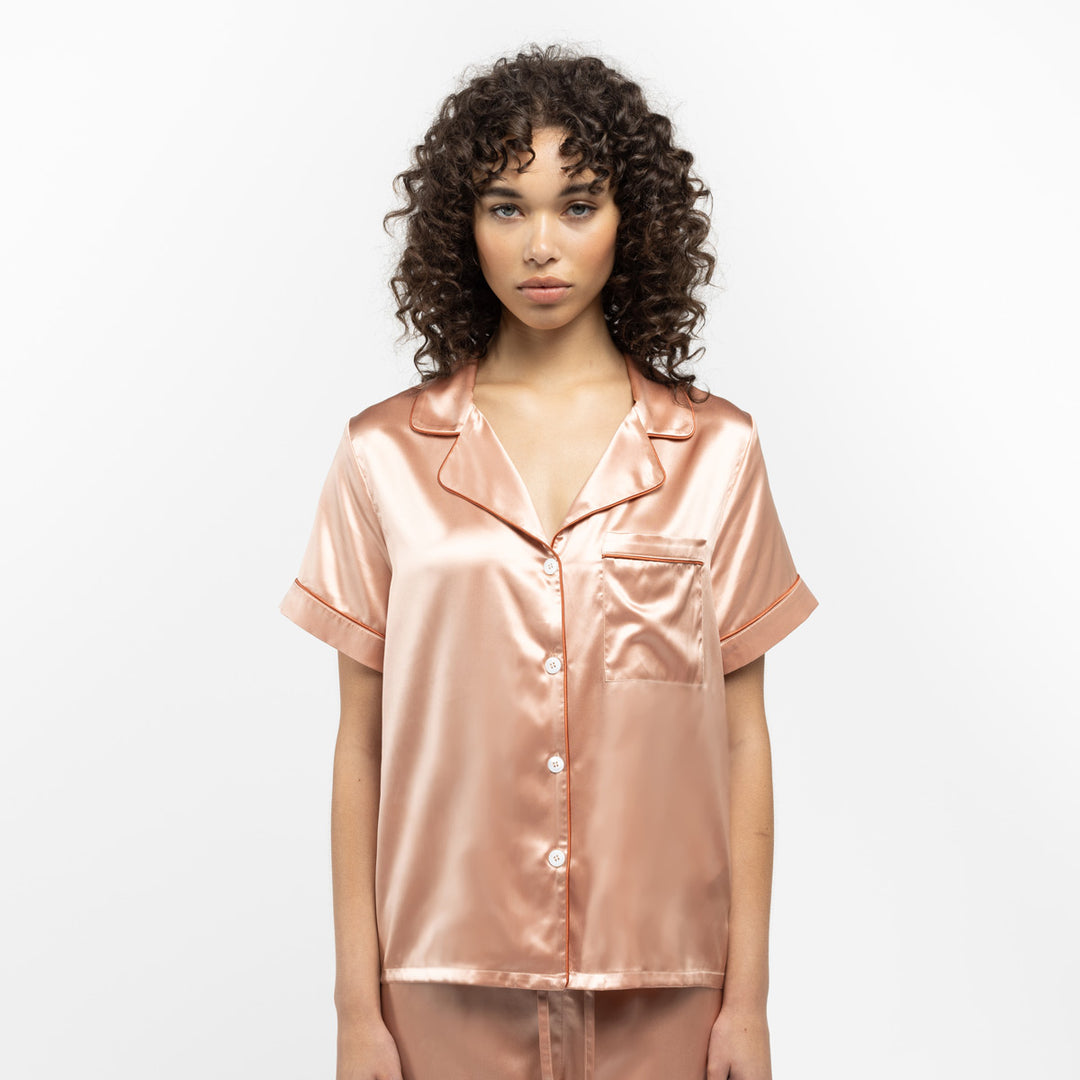 Woman wearing a shiny peach-colored satin pajama top with buttons and a chest pocket#color_clementine-peach-and-dusk