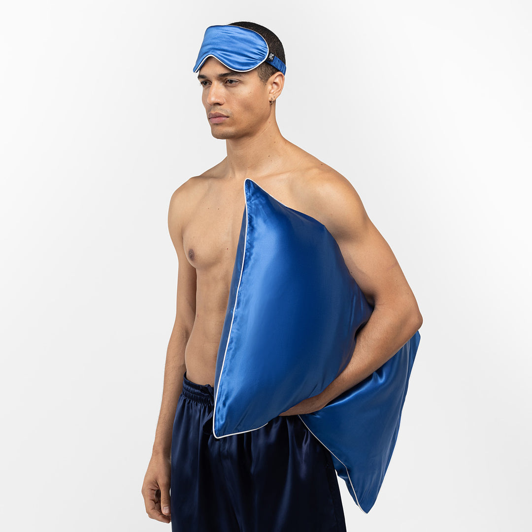 Shirtless man wearing a blue silk sleep mask and navy silk shorts, holding a matching blue silk pillow. - #color_ocean