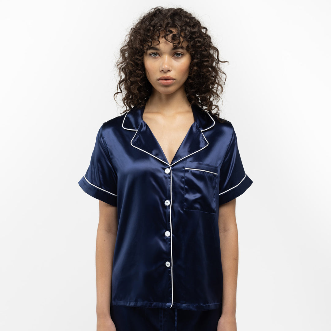 Woman wearing navy blue satin pajamas with white piping, standing against a white background.#color_navy-blue-and-white