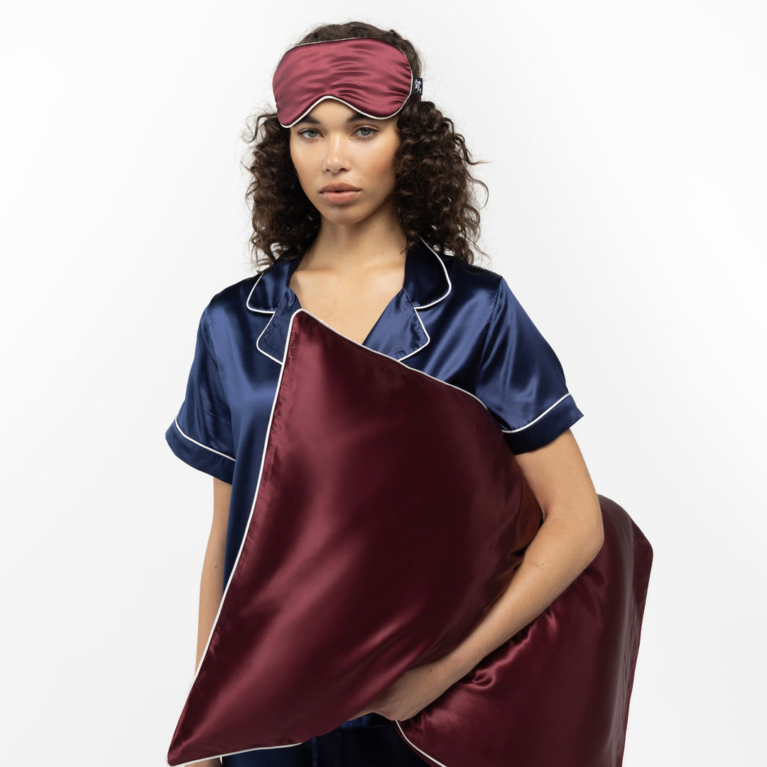 Woman in navy silk pajamas holding a plum silk pillow and wearing a matching plum sleep mask - #color_plum