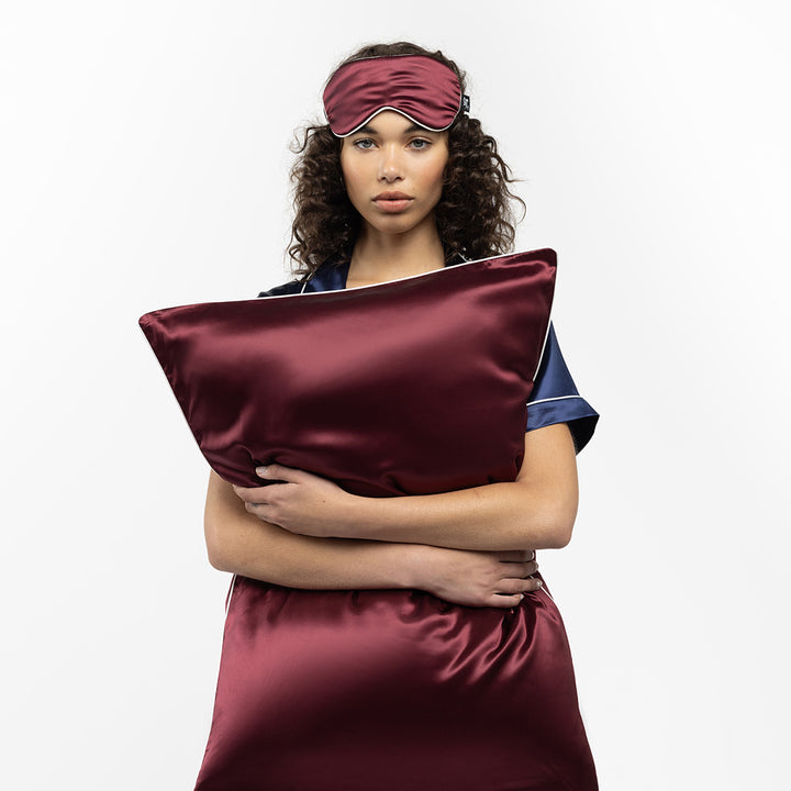 Woman holding a plum-colored silk pillow and wearing a matching sleep mask - #color_plum