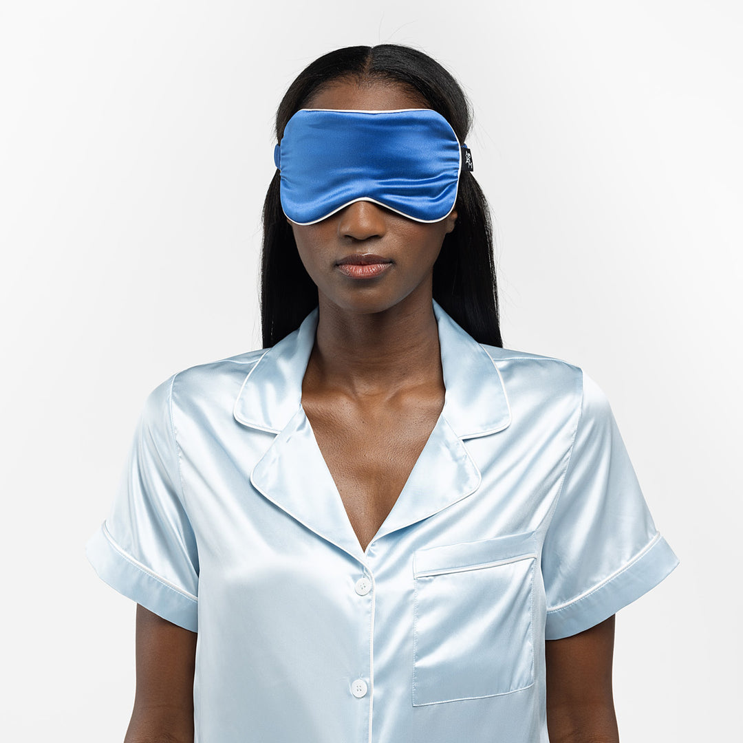 Woman wearing a blue sleep mask and light blue silk pajamas against a white background - #color_ocean