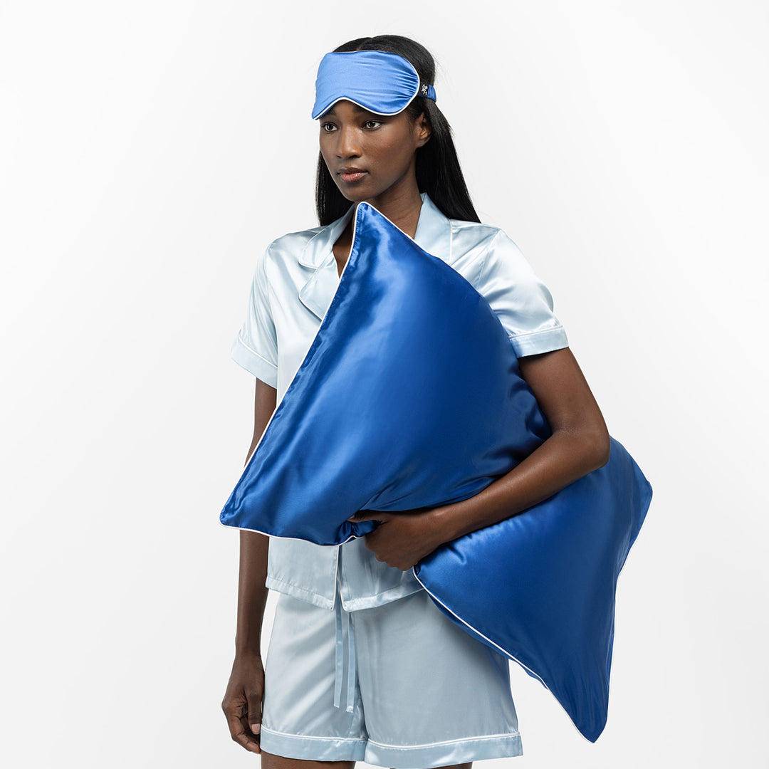 Woman in light-blue silk pajamas holding two deep-blue silk pillows, wearing blue sleep mask - #color_ocean