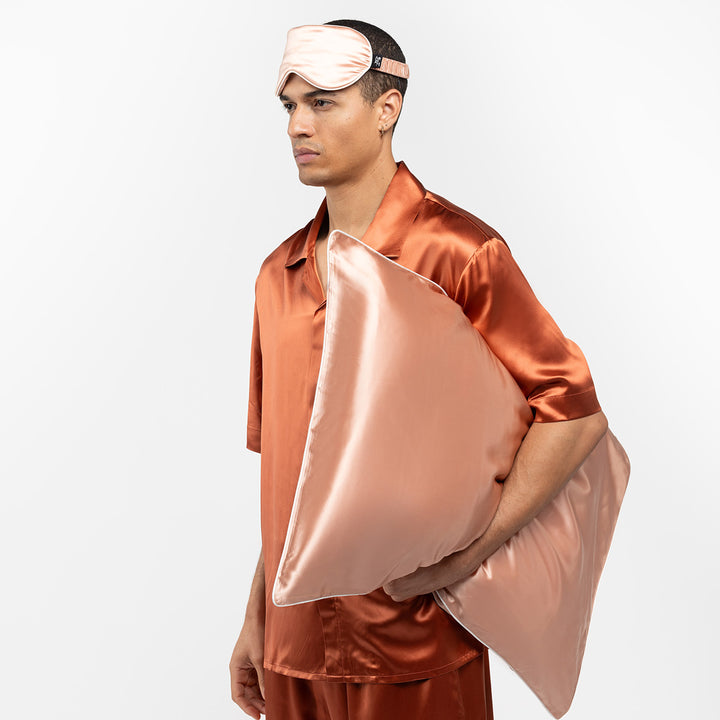 Man wearing clementine-peach silk pajamas and sleep mask, holding a matching silk pillow. - #color_clementine-peach