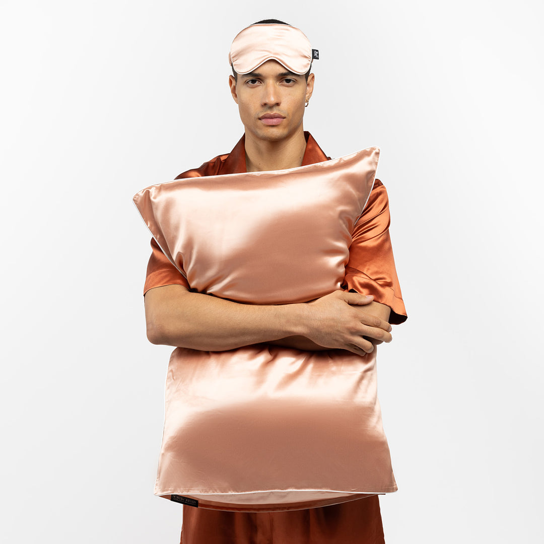Man wearing peach silk pajamas and sleep mask, hugging a matching peach silk pillow. - #color_clementine-peach