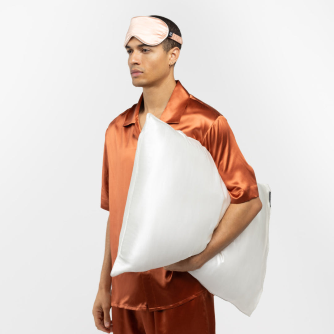 Man in copper silk pajamas wearing a pearl-white silk sleep mask and holding two large white pillows - #color_pearl-white