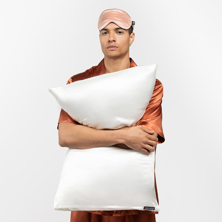 Man in rust silk pajamas wearing a pink sleep mask, hugging two pearl-white silk pillows. - #color_pearl-white