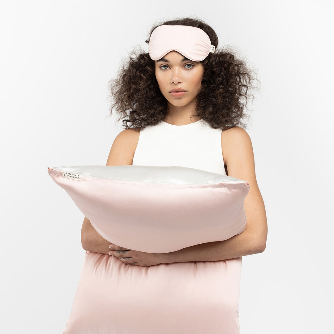 Woman holding a ballet pink pillow and wearing a matching sleep mask, tag reads 'ESSENTIALS BY CLEMENTINE' - #color_ballet-pink