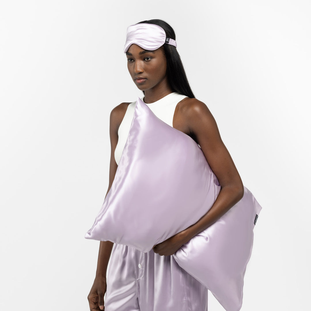 Woman holding a lilac silk pillow and wearing a matching lilac silk sleep mask - #color_lilac-haze