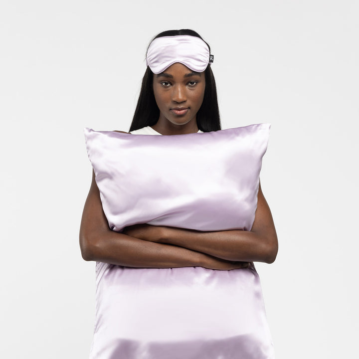 Woman wearing lilac silk sleep mask, arms crossed holding matching lilac silk pillow and duvet - organic pillow high fashion#color_lilac-haze