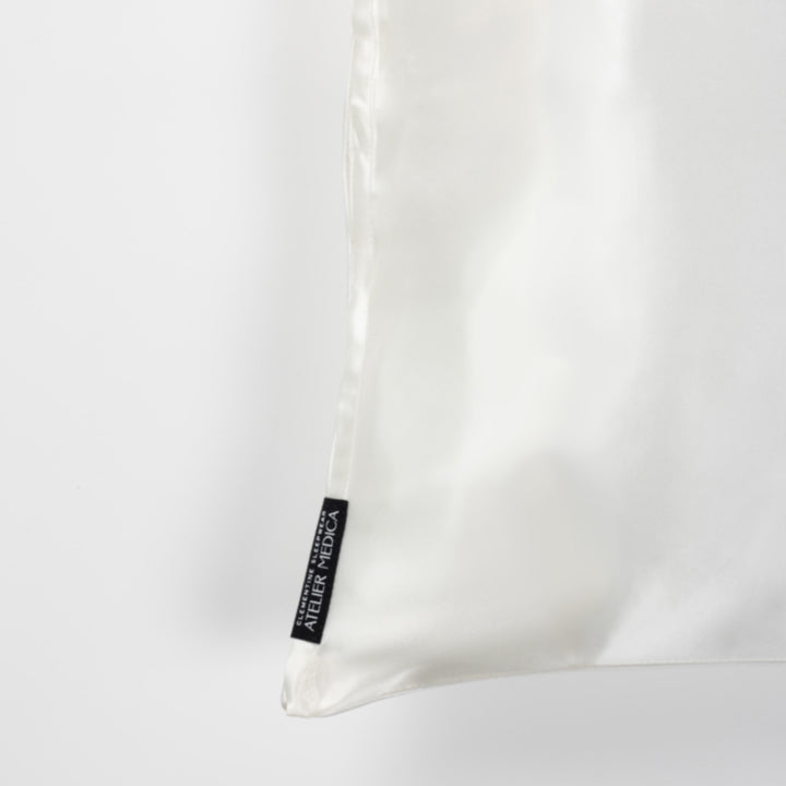 Pearl-white silk pillowcase corner with black brand tag reading 'ATELIER MEDICA'. - #color_pearl-white