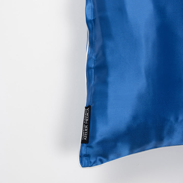 Blue silk pillow corner with white piping and black brand tag reading 'ATELIER MEDICA'. - #color_ocean