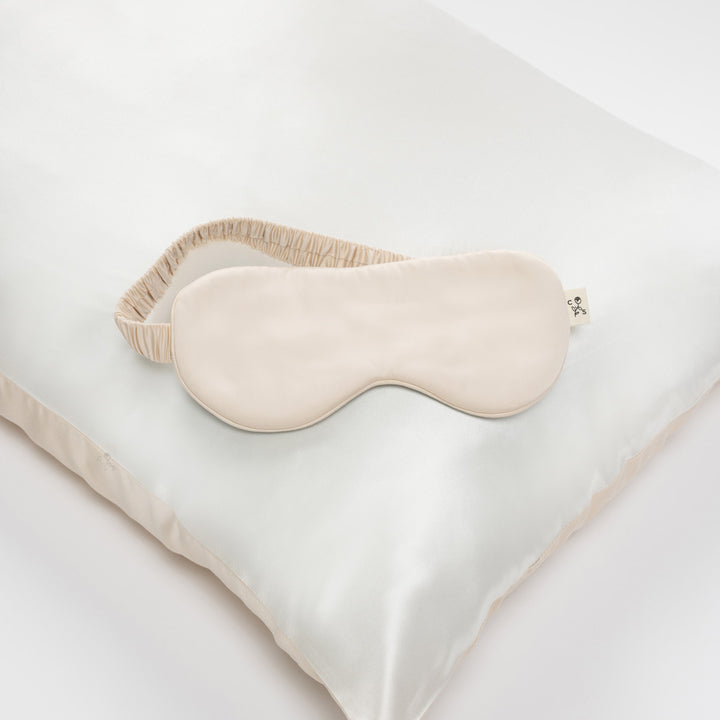 Champagne silk sleep mask with ruched elastic strap resting on a light silk pillowcase - #color_champagne-sunset