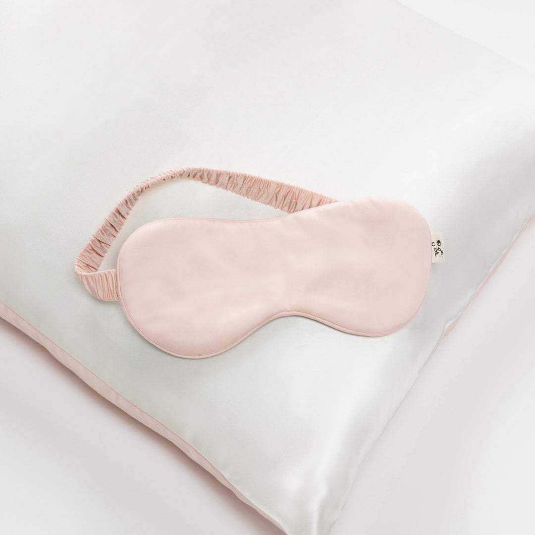 Ballet-pink silk sleep mask with ruched elastic strap resting on a white silk pillow - #color_ballet-pink