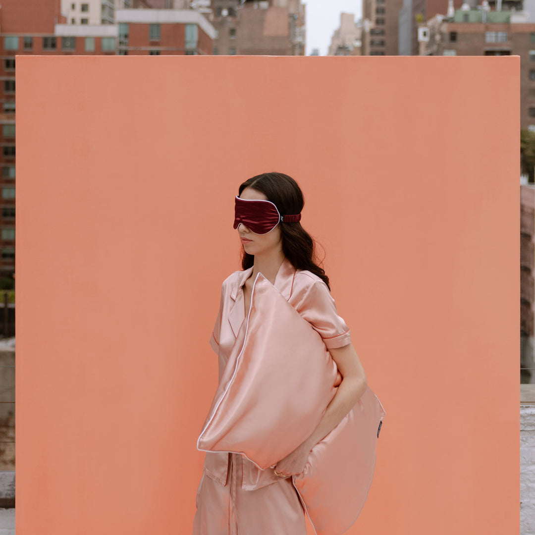 Woman in peach silk pajamas holding a matching pillow, wearing a burgundy sleep mask against a clementine-peach backdrop - #color_clementine-peach