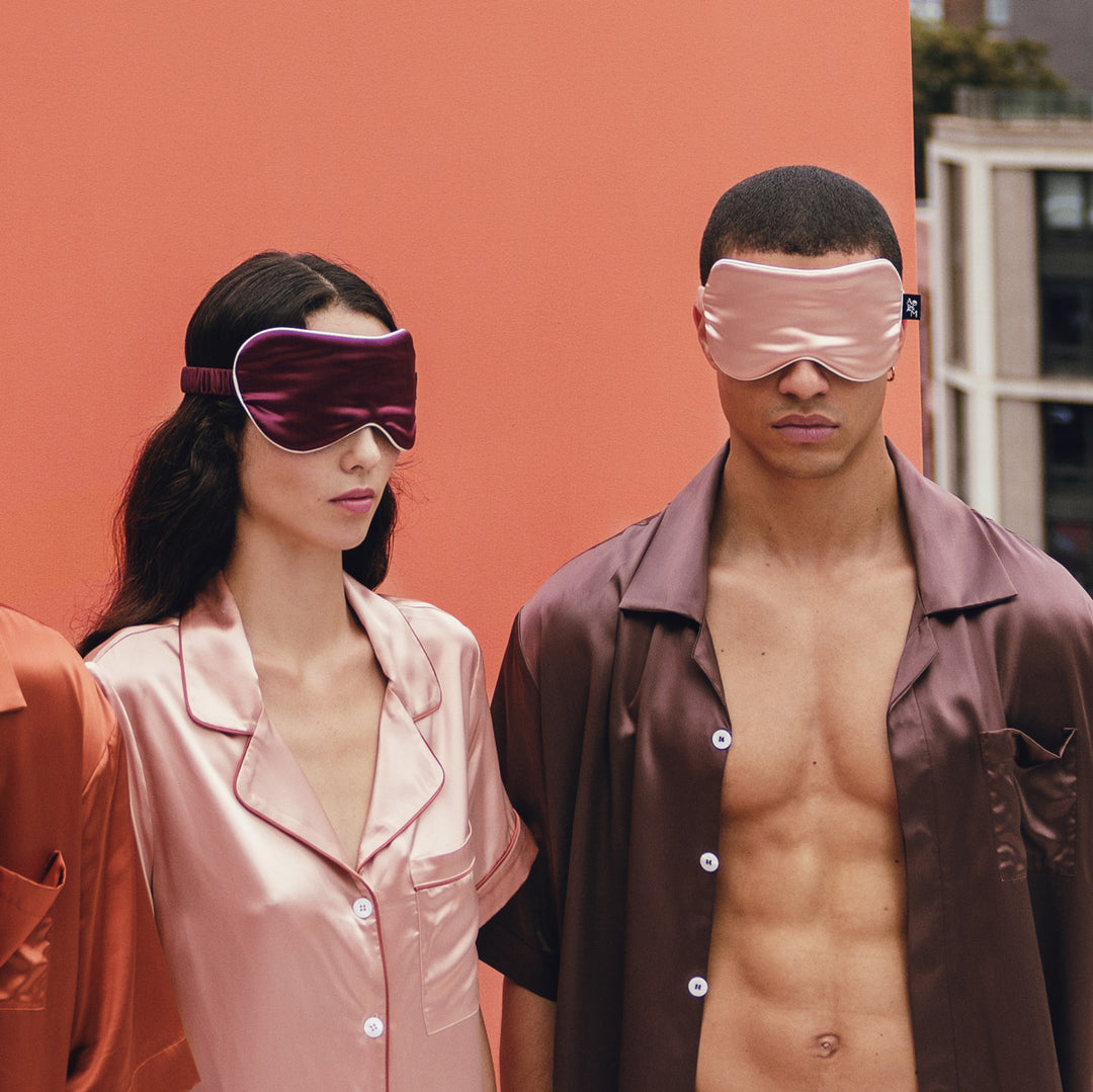 Two people wearing silk sleep masks and pajamas against a peach-colored background - #color_clementine-peach