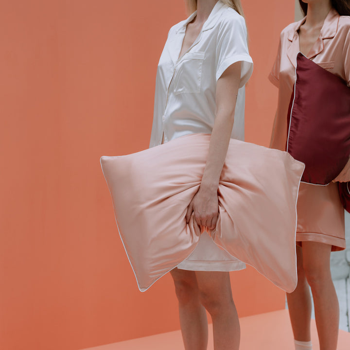 Two women in silk pajamas holding blush and burgundy pillows against a clementine-peach background. - #color_clementine-peach