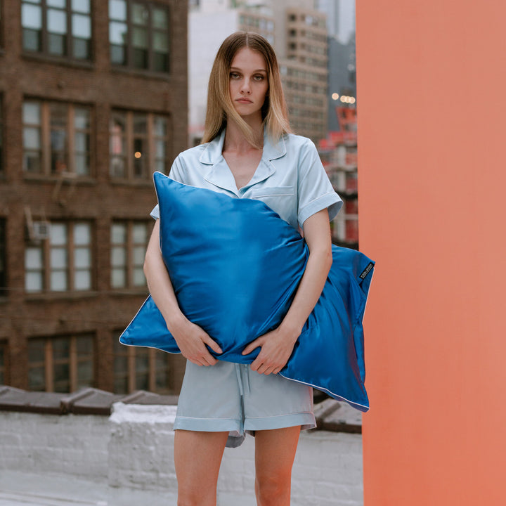 Woman in light blue pajamas holding a bright blue pillow outdoors with buildings in background - #color_ocean