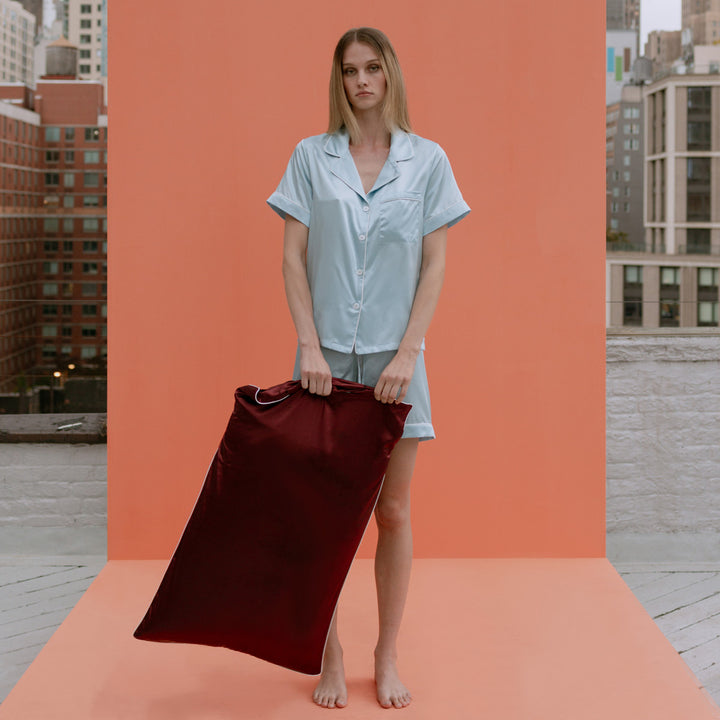 Woman in light blue silk pajamas holding large plum-colored silk pillow against peach backdrop on rooftop - #color_plum