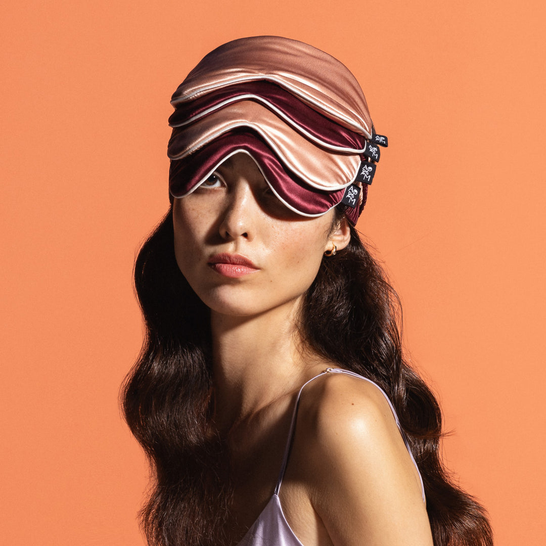 Woman wearing multiple plum and peach colored sleep masks stacked on her head against an orange background - #color_plum