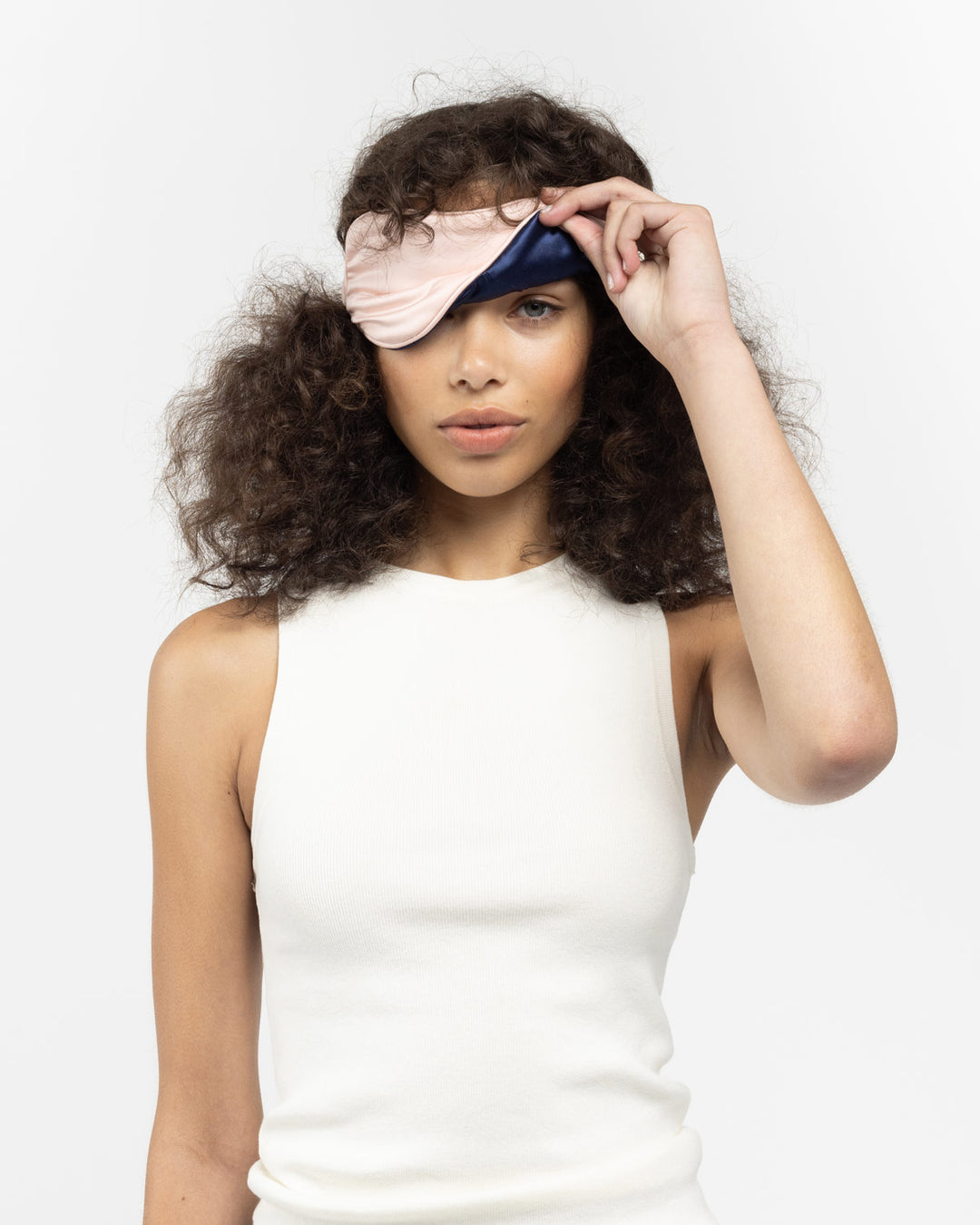 Model lifting a pink-and-navy silk sleep mask to reveal one eye